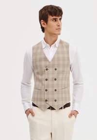 Beżowa kamizelka w kratę z podwójnym zapięciem, brązowymi guzikami, dwoma kieszeniami i ostrym kołnierzykiem, nałożona na białą koszulę.