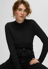 Juodas tekstūruotas viršus su aukštu kaklu ir ilgomis rankovėmis, pagamintas iš raštuotos medžiagos. Dėvima su aukštu taliju juodomis džinsinėmis kelnėmis.