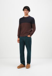 Jeune homme portant un pull côtelé bicolore marron et marine, un pantalon vert foncé et des chaussures à lacets couleur beige, se tenant devant un fond blanc.
