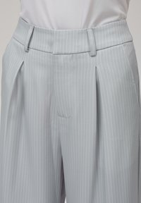 Lys grå bukser med vertikale hvite striper, med flat front, beltehemper og sidelommer. Stoffet har en jevn tekstur.