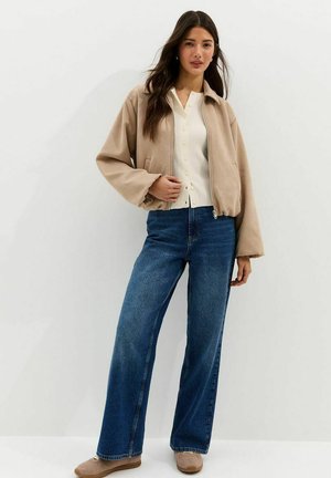 Mujer con chaqueta beige con cremallera, camisa blanca abotonada, vaqueros de pierna ancha azules y zapatillas beige, de pie contra un fondo blanco.