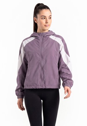 Frau trägt eine lila Kapuzenjacke mit Reißverschluss und weißen Streifen an den Ärmeln sowie schwarze Leggings und schaut leicht nach rechts.