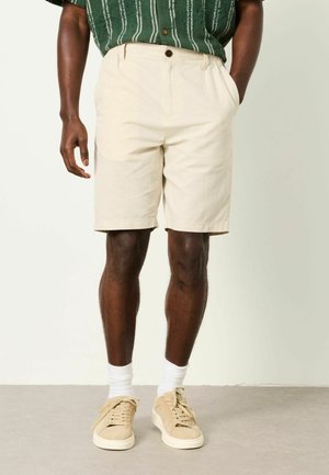 Man wearing beige knee-length shorts with side pockets, a green striped shirt, white socks, and beige sneakers, standing against a plain background.