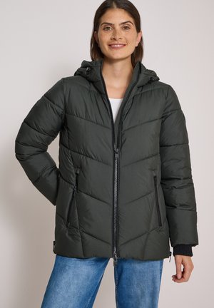 Femme portant une veste matelassée noire à capuche avec poches zippées et jean bleu, souriante, une main sur la hanche, sur un fond uni.