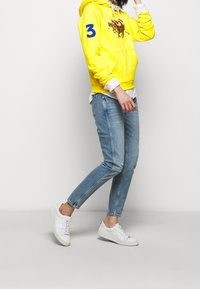 Sudadera amarilla con un diseño gráfico y el número "3" en azul en la manga, combinada con jeans claros desgastados y zapatillas blancas.