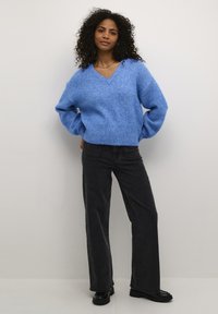 Lichtblauwe gebreide trui met een V-hals en losse mouwen, gecombineerd met zwarte wijde jeans en zwarte schoenen. Soepele textuur.