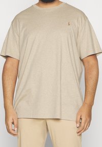 Beige oversize tričko na osobě s krátkými tmavými vlasy a vousy, stojící proti jednoduchému bílému pozadí.