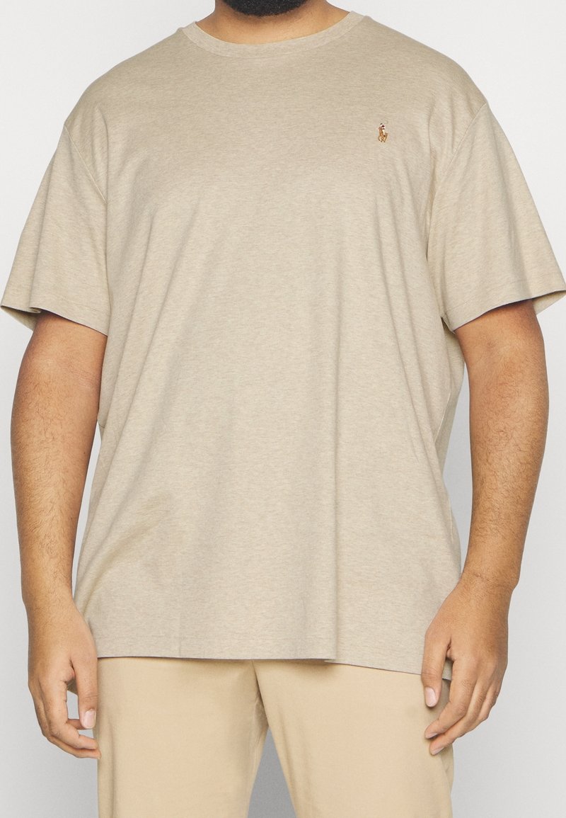 Beige oversize tričko na osobě s krátkými tmavými vlasy a vousy, stojící proti jednoduchému bílému pozadí.