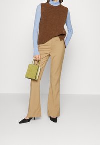 Top sin mangas de punto marrón sobre una camisa de manga larga azul claro, combinado con pantalones acampanados beige y un pequeño bolso texturizado verde.