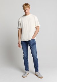 Crème T-shirt met korte mouwen, gecombineerd met blauwe denim jeans. De T-shirt heeft een relaxte pasvorm; de jeans zijn iets tapered. Grijze sneakers met tan zolen.