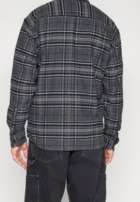 Camisa a cuadros en negro y gris con mangas largas, puños abotonados y un diseño de espalda recta, que presenta una tela suave y texturizada.