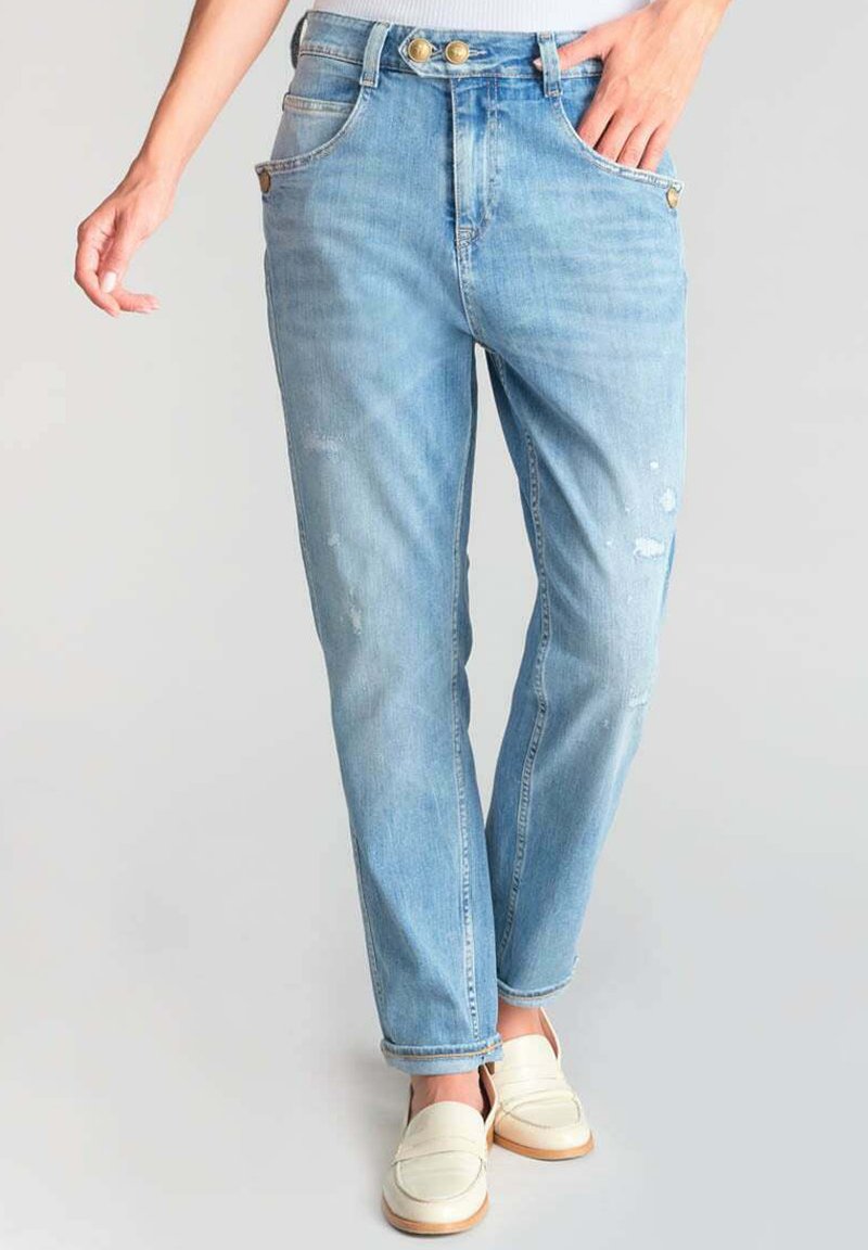 Jean en denim bleu clair à taille haute, avec deux boutons dorés, des patches usés et des jambes droites. Portés avec des mocassins blancs.