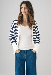 Cardigan a righe in navy e bianco, con una morbida texture a maglia, apertura frontale e polsini a costine. Indossato sopra una canotta bianca.