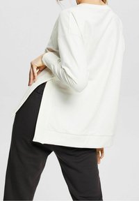 Top blanco ligero de manga larga con aberturas laterales, dobladillo redondeado y una textura suave, combinado con pantalones oscuros.