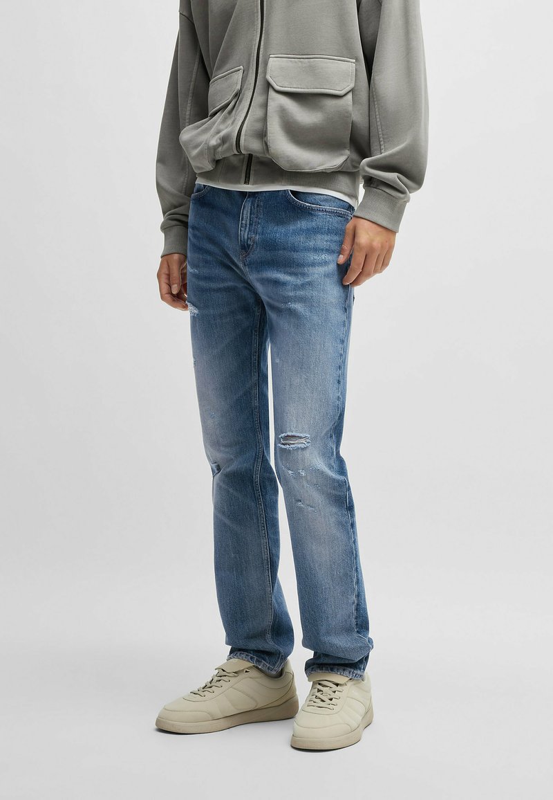 Ljusblå denimjeans med rak passform, prydda med slitna knädetaljer, tillsammans med krämfärgade sneakers och en grå hoodie.