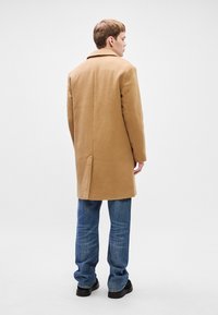 Κάπα από καφέ μαλλί με έναν μόνο πίσω αεραγωγό, κλασικό γιακά με γωνίες και ευθεία κοπή, σε συνδυασμό με μπλε τζιν και μαύρα παπούτσια.