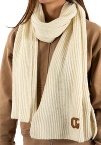 Écharpe en maille côtelée crème avec une texture douce, ornée d'un accent en cuir portant un logo, portée sur un hoodie zippé marron.