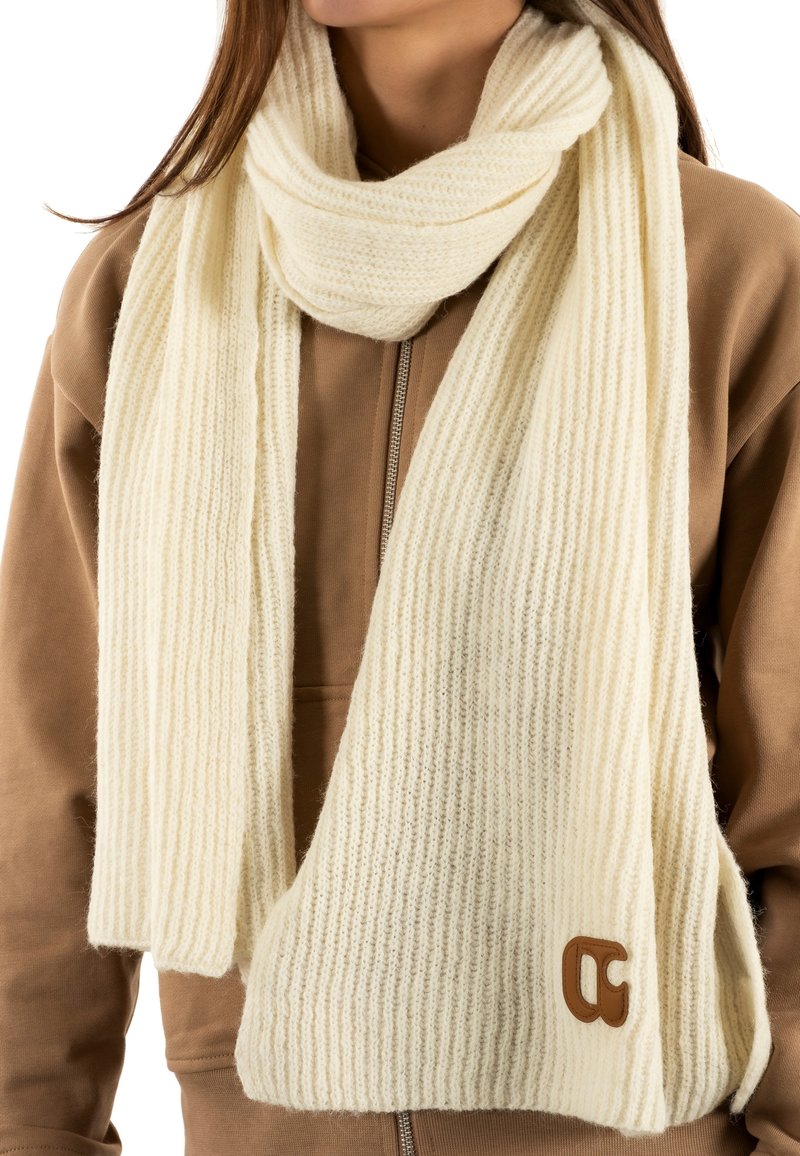 Écharpe en maille côtelée crème avec une texture douce, ornée d'un accent en cuir portant un logo, portée sur un hoodie zippé marron.