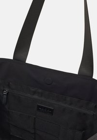 Sort stof tote tas med en top lynlås og frontlommer. Har tekstureret materiale, brede stropper og en cirkulær detalje.