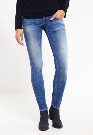 Persoon draagt vervaagde blauwe skinny jeans, zwarte enkellaarzen en een zwarte trui, staand met één hand in de zak.