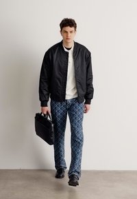 Chaqueta bomber negra, camisa blanca, pantalones azules con patrones geométricos, llevando una bolsa negra. Texturas suaves y ajuste estructurado.