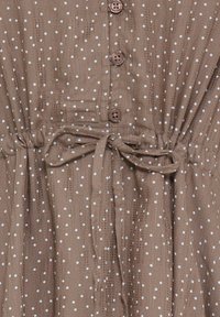 Tissu marron à pois blancs, quatre boutons sur le devant, et une ceinture nouée à la taille sur un matériau texturé.