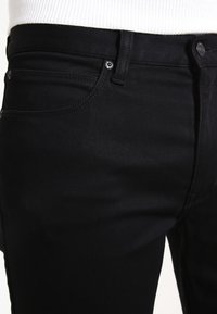 Jeans de denim negro con un diseño ajustado, que presentan bolsillos delanteros, pasadores para cinturón y un cierre de botón brillante. Textura suave y sin patrones visibles.
