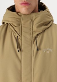 Khaki gekleed jasje met capuchon, gemaakt van gladde stof met een hoge kraag, voorzien van zwarte trekkoorden en geborduurde branding op de borst.