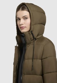 Olivengrøn pufferjakke med hætte, der har lynlås foran og snore, quiltet tekstur og en sort mærkeetiket langs lynlåsen.