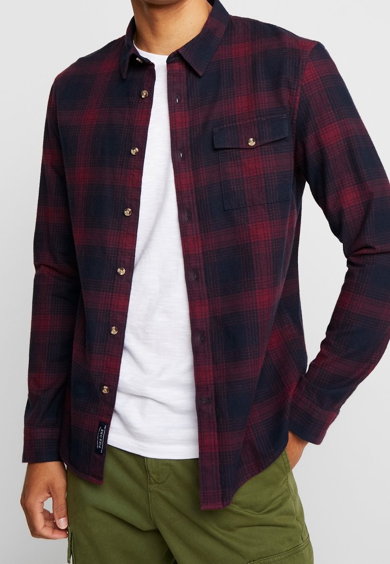 Camicia in flanella a quadri rossi e blu con maniche lunghe, chiusura a bottoni, taschino sul petto e bottoni dorati. Indossata sopra una maglietta bianca.