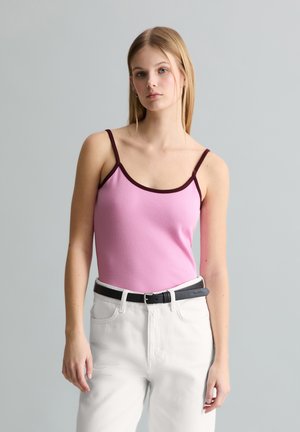Jeune femme portant un top à fines bretelles rose avec une bordure sombre et un pantalon blanc taille haute avec une ceinture noire, debout devant un fond gris.