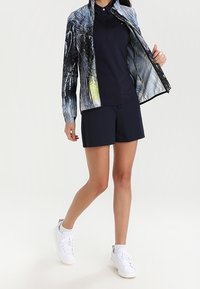 Marinblå shorts i kombination med en matchande polo och en zip-up jacka med ett flerfärgat abstrakt mönster. Vita sneakers fulländar stilen.