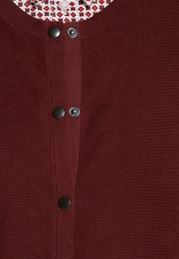 Cardigan bordeaux con un maglione testurizzato, caratterizzato da bottoni a pressione neri e un colletto bianco a fantasia visibile sotto.