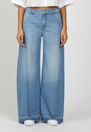 Wide-leg light blue denim jeans featuring a button-fly closure, two front pockets, and a frayed hem. Styled with white shoes.