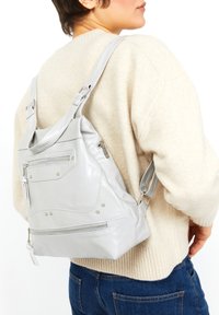 Mochila de cuero gris con una textura brillante, múltiples bolsillos con cremallera y acentos de tachuelas. Con correas ajustables, forma desenfadada y diseño moderno.