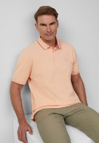 Helles Pfirsich-Polohemd mit einem kontrastierenden burgunderfarbenen Kragen, zwei-Knopf-Leiste und gesticktem Logo. Getragen mit Khakihosen, sitzend.