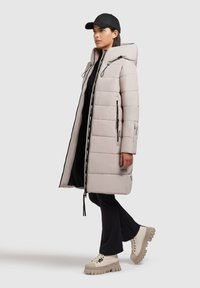 Manteau long rembourré en beige clair avec capuche, double fermeture éclair et poches latérales. Associé à un pantalon noir et des baskets épaisses de couleur claire.