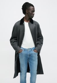 Gara, tumši pelēka virsjaka ar melnu apkakli, valkāta pār baltu kreklu un gaiši ziliem džinsiem. Virsjakai ir aizdare ar sprādzēm un brīvs piegriezums.