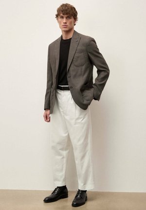 Mlad moški, oblečen v rjavo blazer jakno, črno srajco, bele hlače in črne čevlje, stoji z eno roko v žepu pred enobarvnim ozadjem.