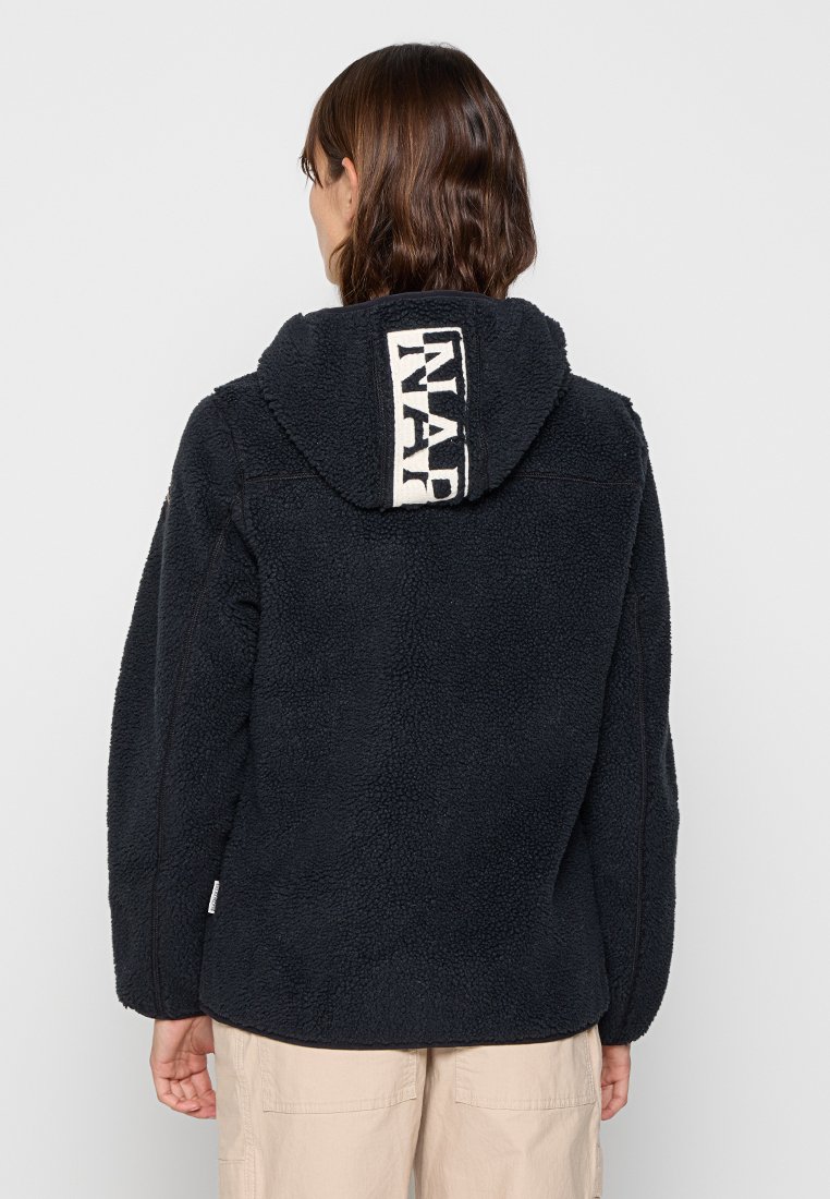 Personne portant une veste en polaire noire texturée avec capuche, affichant un logo vertical blanc à l'arrière, associée à un pantalon beige clair.