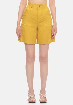 Donna che indossa pantaloncini giallo senape a vita alta con tasche laterali e bottone, abbinati a sandali bianchi con cinturini, in piedi davanti a uno sfondo bianco.