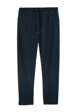 Uomo con giacca bomber nera, felpa blu navy, pantaloni blu navy con coulisse e sneakers slip-on nere con suola bianca, in piedi con le mani in tasca.