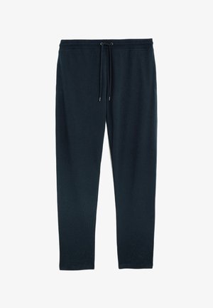 Homme portant une veste bomber noire, un sweat-shirt bleu marine, un pantalon bleu marine à cordon de serrage et des baskets noires faciles à enfiler avec des semelles blanches, debout les mains dans les poches.