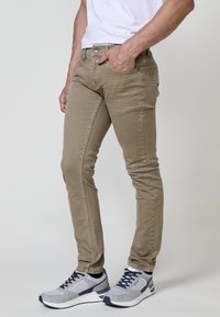 Khaki skinny farmer nadrág diszkrét mintázattal, elülső zsebekkel és fém gombos záródással. Szürke sportcipővel párosítva, kék részletekkel.