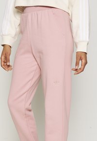 Person som bär ljusrosa Adidas-träningsbyxor och en krämfärgad långärmad tröja med vita ränder på ärmarna.