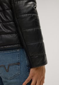 Schwarze Lederjacke mit gestepptem Muster, ausgestattet mit gepolsterten Abschnitten und einer glatten Oberfläche, kombiniert mit hellblauen Jeans.