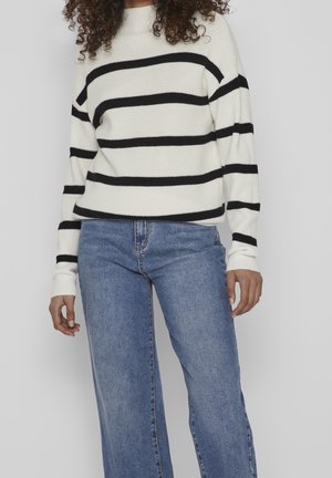 Personne aux cheveux bouclés portant un pull blanc à rayures horizontales noires et un jean bleu taille haute sur un fond gris uni.