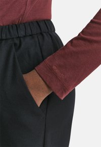 Hand in der Tasche von schwarzen elastischen Hosen mit Gummizug, trägt ein langärmliges burgunderrotes Hemd mit dem aufgenähten GANT-Logo am Ärmel.
