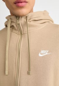 Sweatshirt com fecho éclair Nike bege, com cordões, apresentando um logótipo branco no peito, feito de um suave tecido em mistura de algodão.