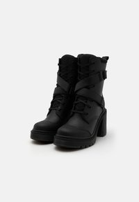 Bottines noires avec un talon épais, dotées d'un laçage à l'avant, de sangles croisées et d'une semelle en caoutchouc texturé. Fabriquées en cuir synthétique.